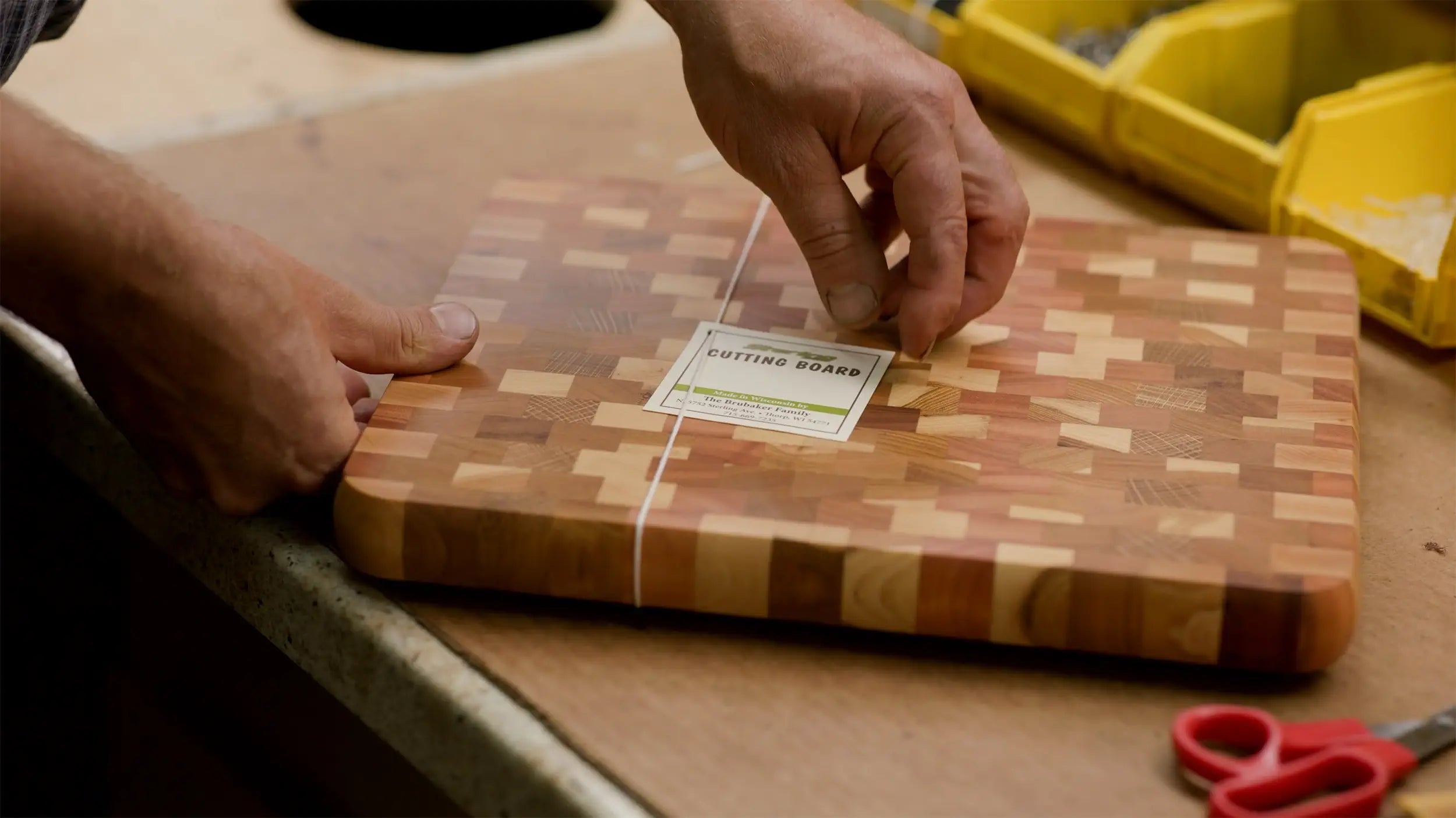 A person placing a label on a wooden end-grain cutting board, with scissors and yellow storage bins in the background.