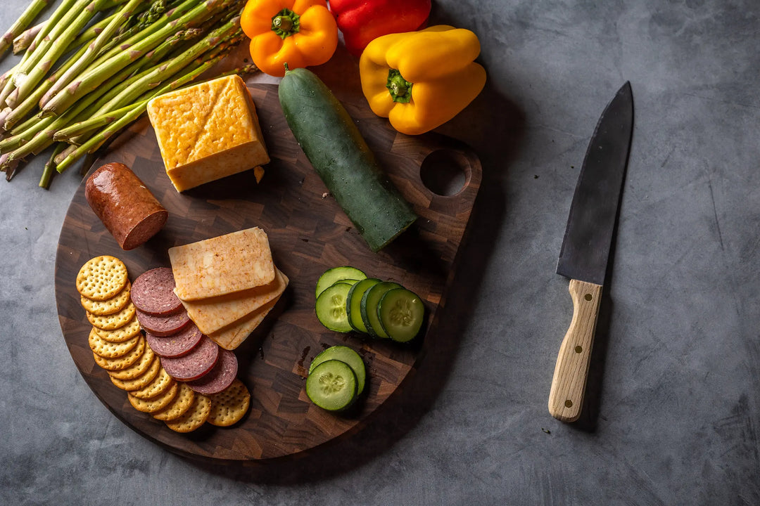 A charcuterie board with sliced cheese, sausage, crackers, cucumber, and whole vegetables like peppers and asparagus, placed next to a knife on a gray surface.