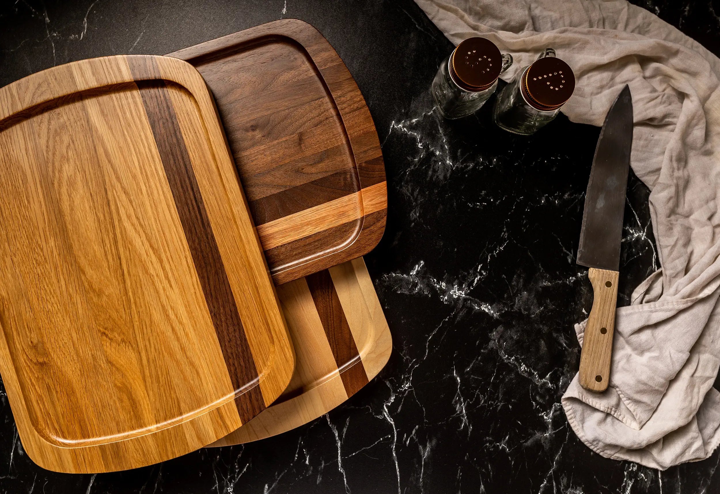 Three wooden cutting boards with different wood tones stacked on a black marble surface, alongside a knife and glass containers with copper lids.