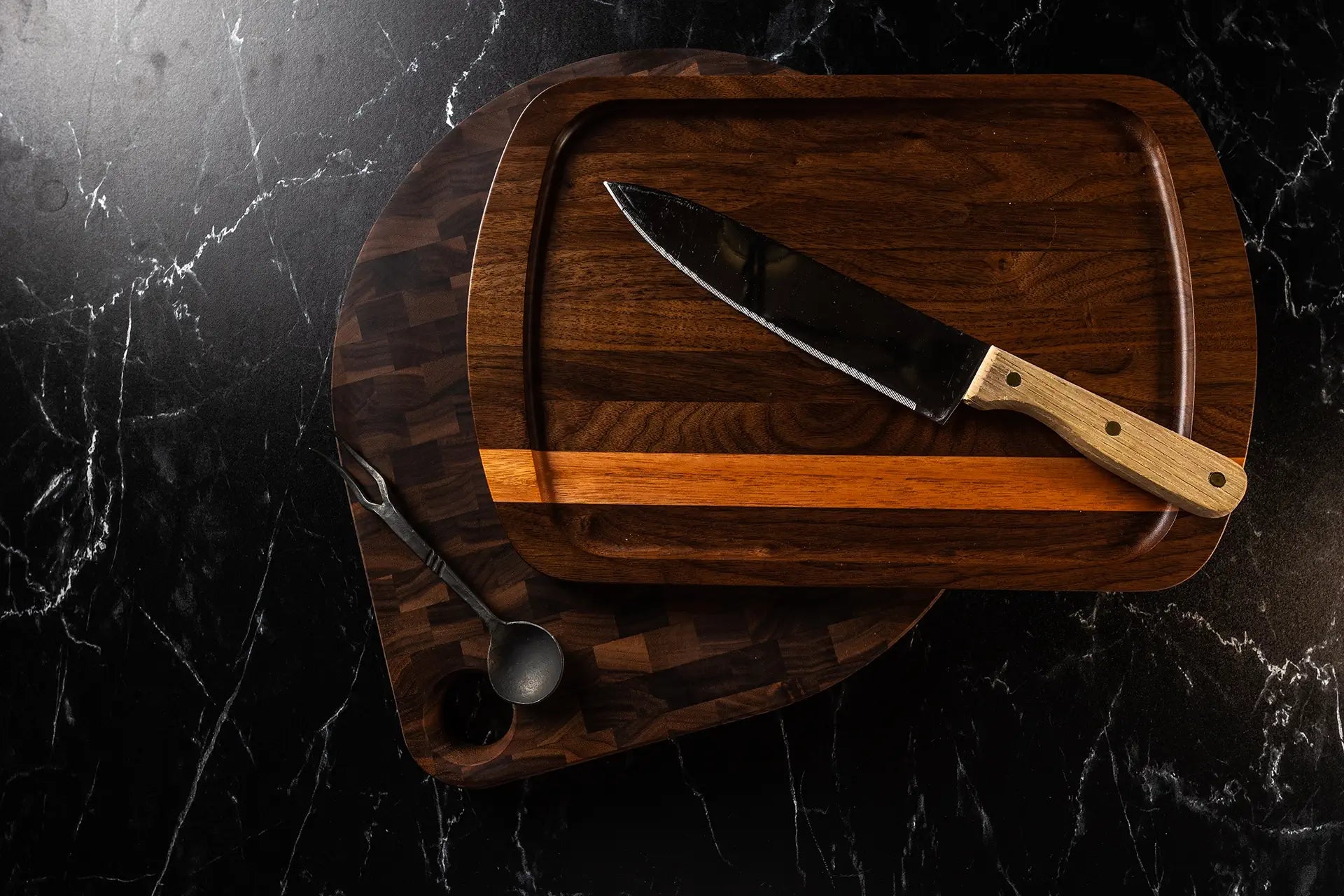 Top view of two dark wooden cutting boards on a black marble countertop, featuring a chef's knife with a wooden handle and a metal serving fork.