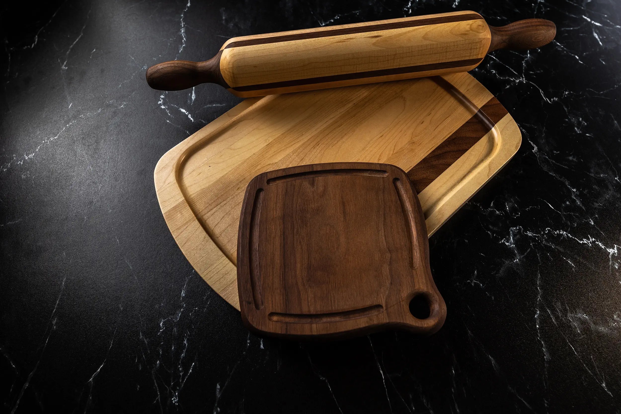 A wooden rolling pin and two cutting boards, one small and one large with a groove, placed on a dark marble countertop.