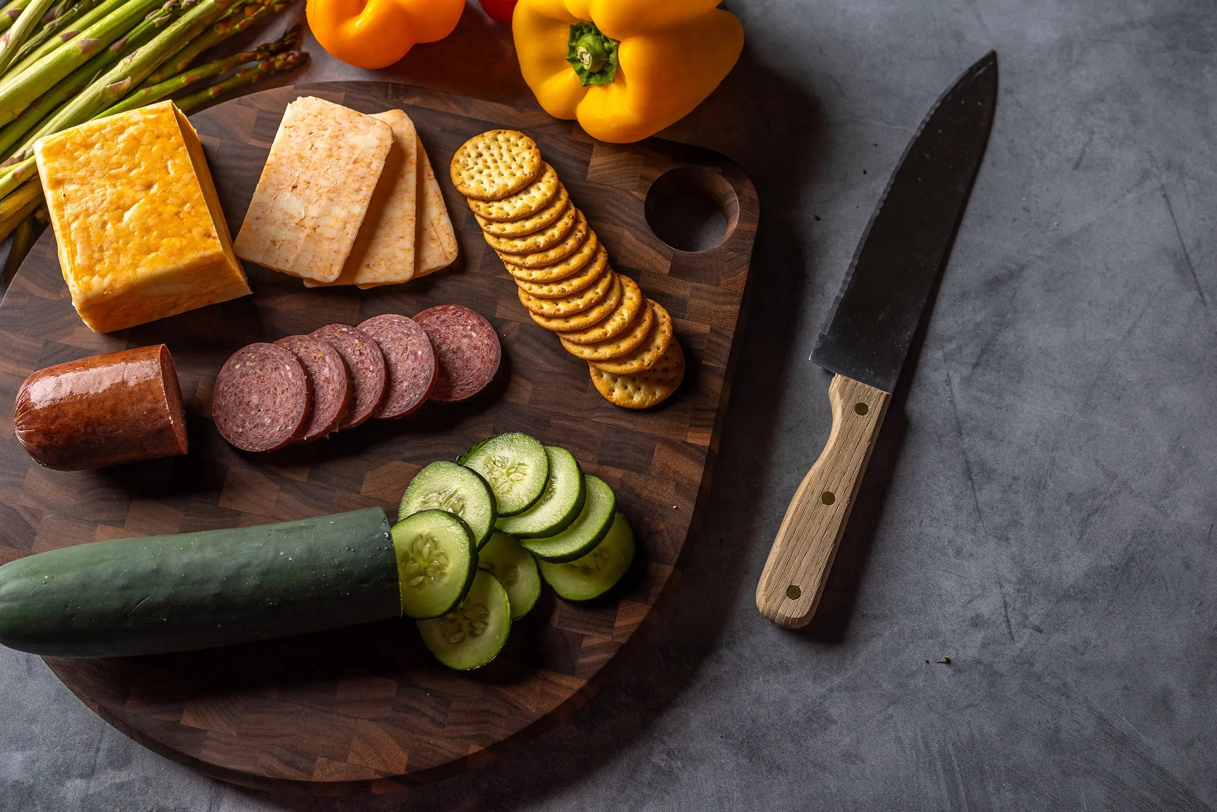 Cutting board with cheese, sausage, cucumber slices, crackers, and vegetables, alongside a chef’s knife.