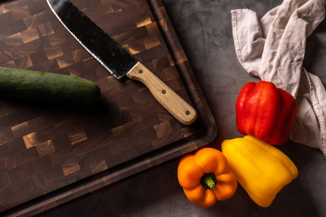 A wooden cutting board with a chef’s knife and cucumber, next to red, yellow, and orange bell peppers on a dark surface with a crumpled cloth nearby.