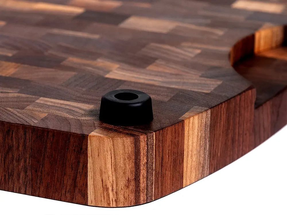 A close-up view of a wooden cutting board with a rich, end-grain pattern and rounded corner details. The board features a black rubber foot for stability, highlighting the craftsmanship and durable design.