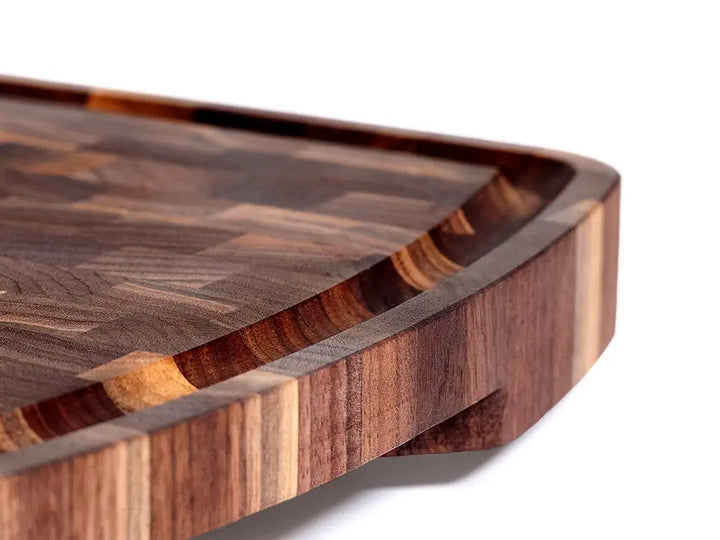 A close-up of a wooden cutting board showcasing a smooth, curved juice groove designed to catch liquids. The board features a rich end-grain pattern with natural wood tones, emphasizing both its functionality and aesthetic craftsmanship.