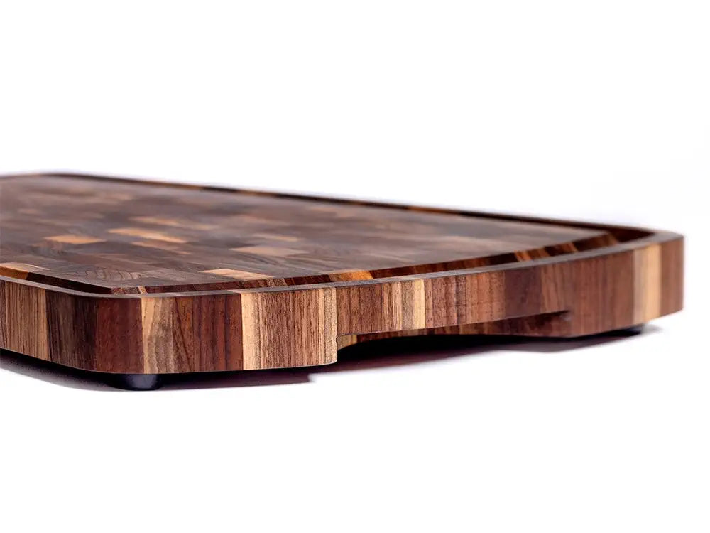 A side-angle view of a wooden cutting board featuring a detailed end-grain pattern, a curved juice groove along the top, and slightly raised black rubber feet. The design highlights both the board’s sturdy construction and elegant wood craftsmanship.