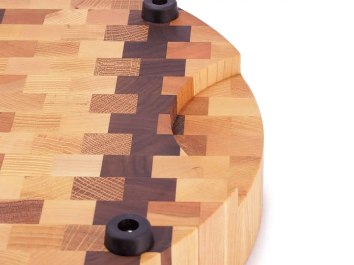 Close-up of a round end grain butcher block with a checkered pattern, showing a handle cutout and black rubber feet, against a white background